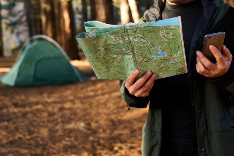 Adult hiker with backpack using mobile phone and map in forest near his tent. Hiking elderly man in autumn nature holding map outdoors next to a campsite in the sunlight 