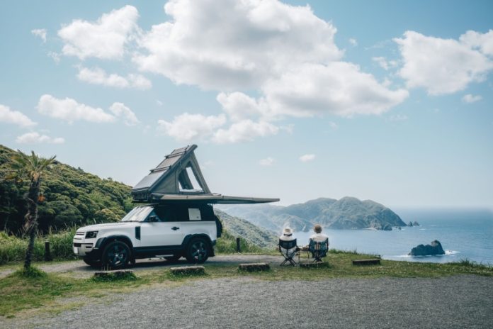 Couple sitting in a camping chair in nature with a view of Island in a beautiful blue sky with an overlanding car with a rooftop tent and an awning.