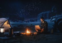 Man sitting by campfire and tent using a laptop while camping